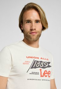 Hombre rubio con barba que lleva una camiseta blanca con el texto y gráficos "Union Alls 1889 Lee Durable Workwear American Original".