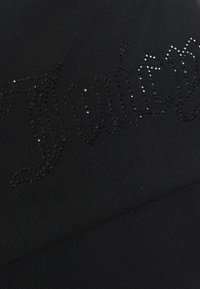 Melna auduma ar gludu tekstūru, kurā ir rhinestone rotājums ar vārdu "Juicy" rakstītu kursīvā fontā.