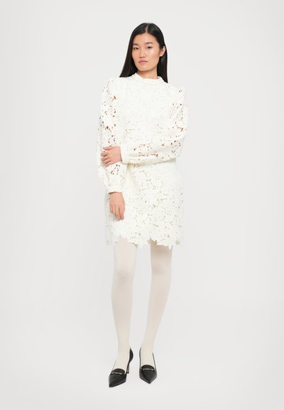 Witte kantjurk met bloemenpatronen, hoge hals en opgezette mouwen, gecombineerd met witte panty’s en zwarte puntige hakken.