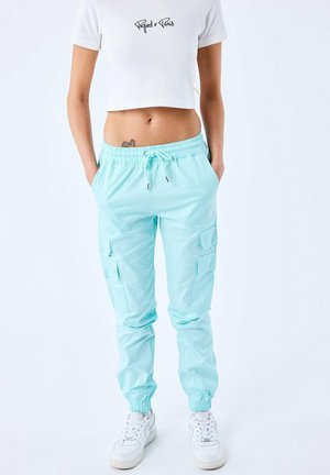 Un pantalon cargo bleu clair avec une taille élastique, un cordon de serrage, des poches latérales et des poignets ajustés. Associé à un haut blanc court.