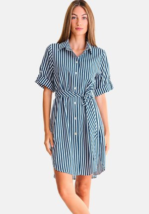 Femme debout portant une robe chemise rayée verticalement bleu et blanc, avec des manches courtes retroussées et une ceinture nouée à la taille.