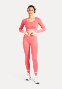 Top corto rosa a maniche lunghe e leggings a vita alta con strisce bianche, realizzati in materiale elasticizzato, abbinati a scarpe da ginnastica bianche.