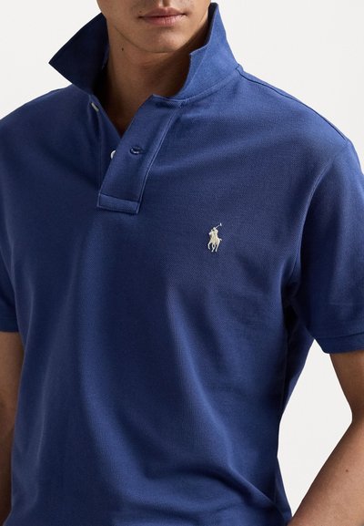 Tumedat sinine polo särk tekstuuriga kudumiga, väljaulatuva krae ja kolmesedme nööbiga. Rinnal on väike valge broderi logo.
