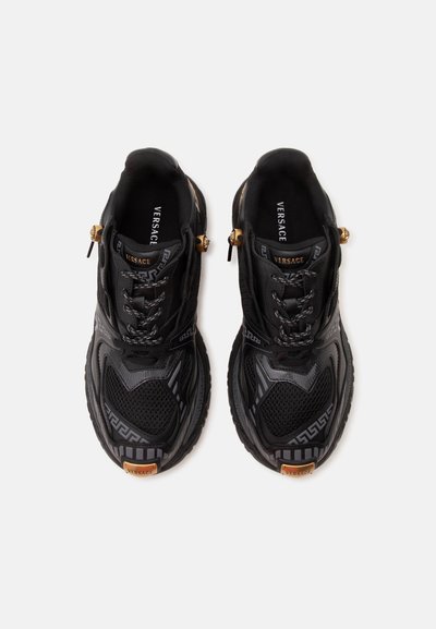 Ζευγάρι μαύρα sneakers Versace με σχεδιασμένο πλέγμα, κορδόνια μπροστά, χρυσές λεπτομέρειες και λογότυπο, σε θέα από πάνω σε λευκό φόντο.