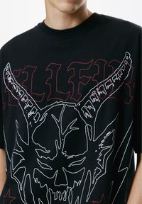 Crna majica s velikim bijelim konturama rogate lubanje i crvenim stiliziranim tekstom djelomično vidljivim iza grafike.