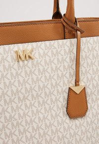 Gros plan sur un sac à main Michael Kors beige et sable avec motif logo MK, initiales MK dorées et étiquette en cuir sable avec accent doré.