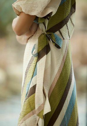 Frau trägt ein strukturiertes Kleid mit diagonalen Streifen in Grün, Blau, Braun und Beige, gebunden zu einer Schleife an der Taille, Arme verschränkt.