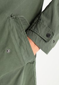 Veste en coton vert avec une poche à rabat et un détail de bouton-pression noir et rond. Le tissu a une finition légèrement texturée.