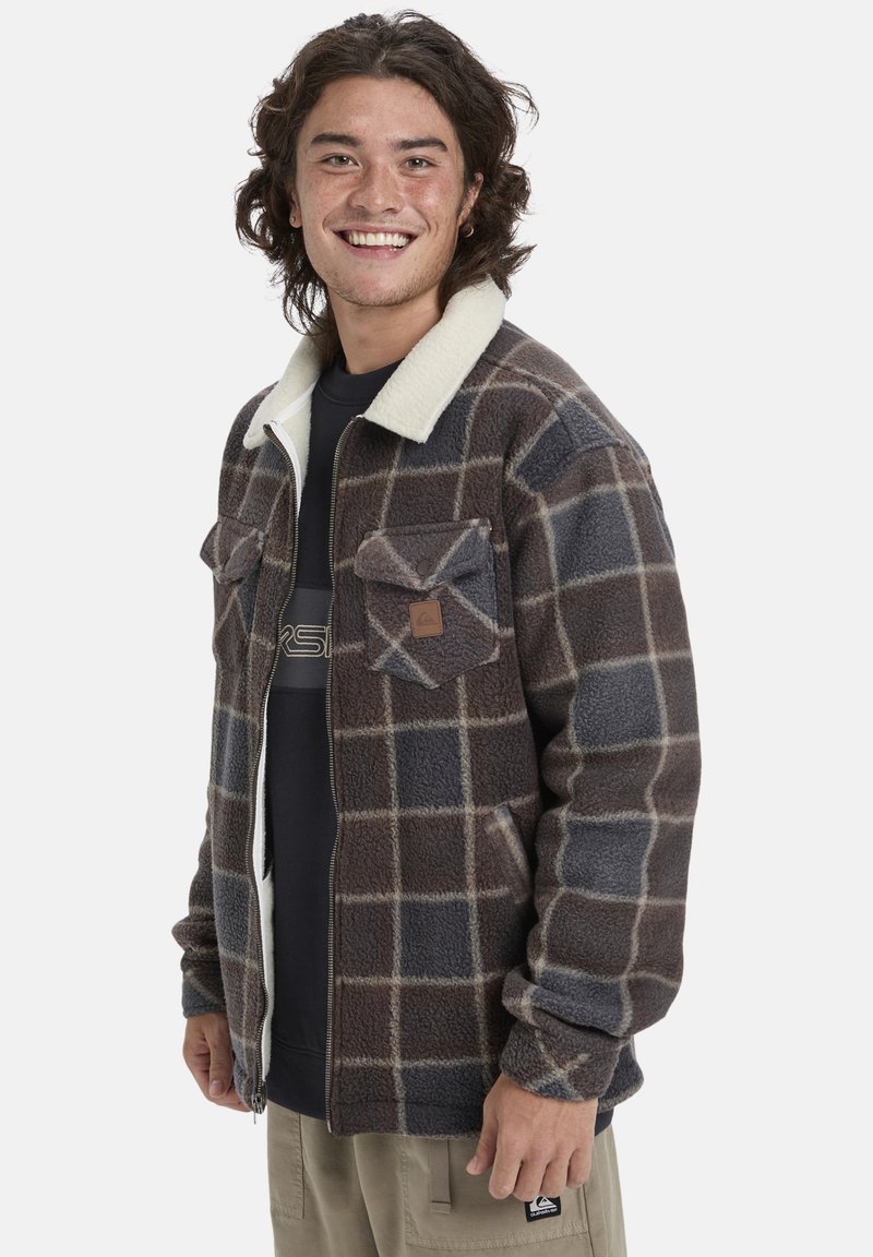 Chaqueta de forro polar con un patrón de cuadros marrón y gris, cuello beige, cierre de cremallera y bolsillos frontales. Interior forrado para mayor calidez y comodidad.