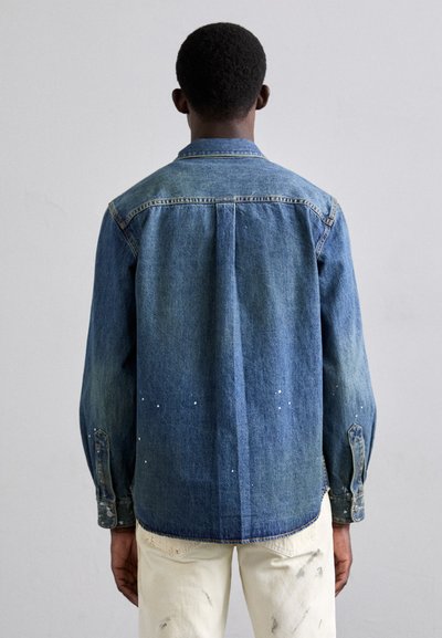 Camisa de mezclilla en varios tonos de azul, con cuello puntiagudo, mangas largas y salpicaduras de pintura blanca dispersas.