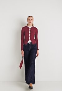 Cardigan burgund cu mâneci lungi și butoane albe, asociat cu blugi negri cu picioare largi și o geantă texturată roșie, completat cu pantofi negri.