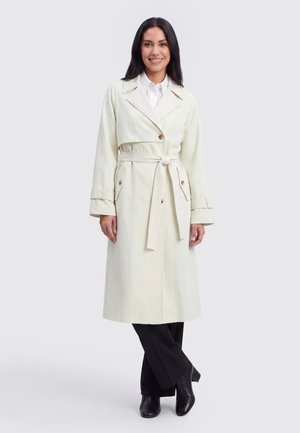 Donna sorridente che indossa un lungo trench color crema con cintura in vita sopra pantaloni neri e stivali neri con tacco, in piedi su uno sfondo semplice.