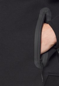 Handen placerad i en vertikal dragkedjeficka med gummiserad kant på ett svart tygplagg.