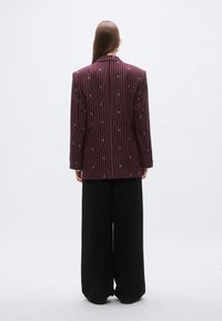 Vino rdeč črtast blazer z navpičnimi črtami in subtilnimi poudarki, kombiniran z ohlapnimi črnimi hlačami, prikazan od zadaj na nevtralnem ozadju.