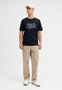 Zwarte crew-neck t-shirt met "Let it snow" graphic, gecombineerd met beige broek en witte sneakers met beige accenten, geleverd in een casual pasvorm.