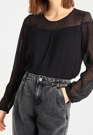 Vrouw draagt een zwarte doorschijnende blouse met lange mouwen en een gestippelde textuur, ingestopt in hooggetailleerde vervaagde zwarte spijkerbroek, één hand in de zak.