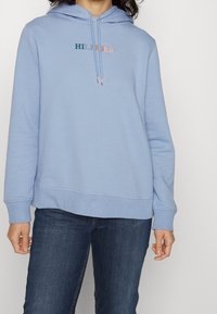 Lichtblauwe katoenen hoodie met trekkoorden, geborduurd "HILFIGER" logo in contrasterende kleuren, en ribgebreide manchetten en zoom. Gecombineerd met denim jeans.