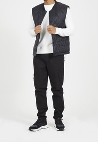 Black padded vest with a quilted pattern, worn over a white long-sleeve shirt, paired with black cargo pants and black sneakers.