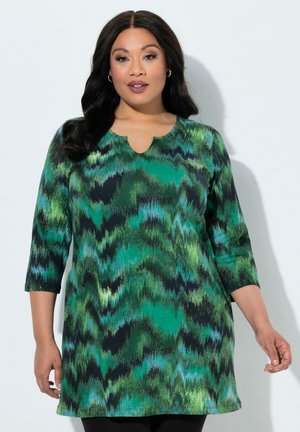 Donna con lunghi capelli scuri indossa un vestito verde e nero a fantasia con maniche a tre quarti, in piedi davanti a uno sfondo chiaro e semplice.