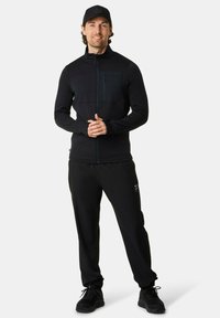 Man in een zwart sportjack, zwarte broek, zwarte sneakers en een zwarte pet, met zijn handen gevouwen staand voor een lichtgrijze achtergrond.