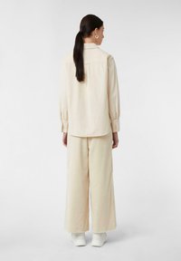 Camicia oversize beige chiaro con maniche lunghe, colletto alto e orlo arrotondato, abbinata a pantaloni a gamba larga e sneakers bianche.