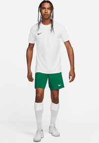 Maglietta Nike bianca a maniche corte con logo verde, abbinata a pantaloni corti verdi con logo Nike bianco. Calze bianche fino al ginocchio e scarpe bianche.