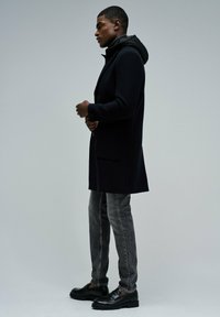 Abrigo negro con capucha, diseño de un solo botón, bolsillos laterales, tejido texturizado; usado sobre jeans grises y mocasines negros.