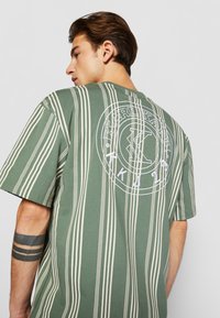 Grönrandig t-shirt med vertikala krämfärgade ränder och en cirkulär vit logotyp på baksidan. Korta ärmar, avslappnad passform, bomullstyg.