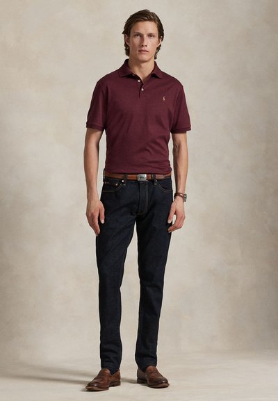 Ung mand stående iført bordeaux polo, mørke jeans, brun bælte, læderur og brune pæne sko mod en neutral baggrund.
