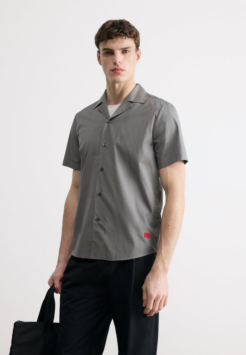 Νεαρός άνδρας που φοράει γκρι κοντομάνικο πουκάμισο με κουμπιά και μαύρο παντελόνι, κρατώντας μια μαύρη τσάντα μπροστά σε απλό λευκό φόντο.