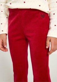 Røde fløjlsbukser med en tekstureret overflade og et lille hjerteemblem på lommen. Kombineret med en hvid top med små hjerter.