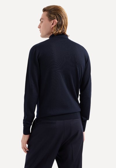 Maglione lavorato a maglia blu marina con colletto alto, polsini e orlo a coste, caratterizzato da un motivo testurizzato. Indossato con pantaloni scuri, visto da dietro.