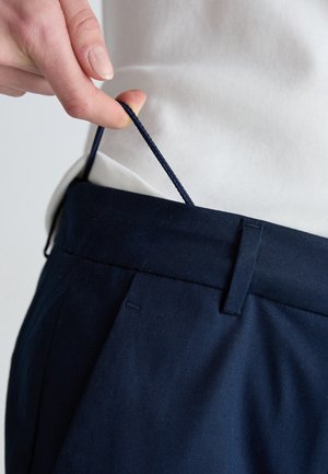 Hånd, der holder justerbar navyblå snor i linningen på navyblå bukser med hvid skjorte stoppet ind.