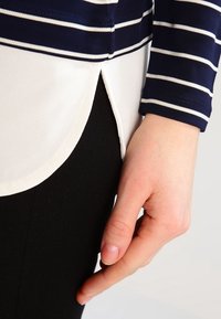 Mörkblå och vit randig långärmad topp med rundad nederkant; tyget är av bomullsblandning. Svarta byxor synliga nedanför. Hand vilande på sidan.