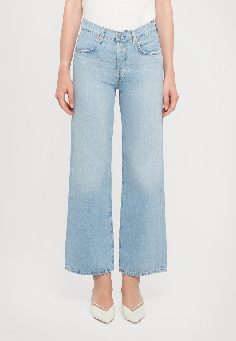 Jeans larghi azzurri realizzati in denim, con vita alta, tasche frontali e orlo pulito. Indossati con scarpe bianche a punta.