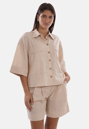 Donna che indossa una camicia beige a maniche corte con bottoni e pantaloncini larghi abbinati, in piedi con una mano in tasca davanti a uno sfondo semplice.