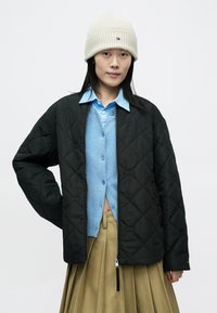 Chaqueta negra acolchada con cierre de cremallera, llevada sobre una camisa azul abotonada y una falda beige, combinada con un gorro de punto color crema.