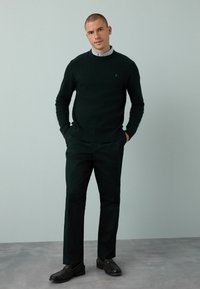 Dunkelgrüner gestrickter Pullover, dunkelgrüne Hose und schwarze Schuhe. Einfaches, strukturiertes Design mit gerippten Bündchen und Kragen, gepflegter Look.