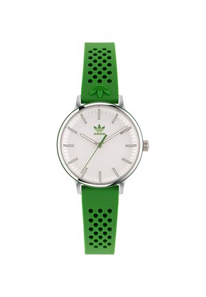 Montre avec bracelet en silicone vert, boîtier rond en argent, cadran blanc et accents argentés. Présente des perforations distinctes sur le bracelet.
