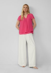 Rosa, kurzärmelige Bluse mit V-Ausschnitt und geschichtetem Saum, kombiniert mit weißen, weiten Hosen. Glatte Stoffqualität, minimale Details.