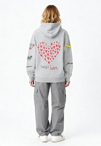Szürke kapucnis pulóver piros szív mintával a hátán, szöveggel és színes grafikákkal díszítve. Szürke terepmintás nadrággal és fehér tornacipővel párosítva.