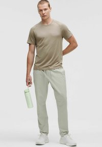 Beige Sport-T-Shirt mit kurzen Ärmeln und hellgrünen Jogginghosen. Das Modell hält eine passende hellgrüne Wasserflasche. Weiße Sneakers.