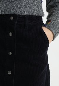 Personne portant une jupe en velours côtelé noire avec des boutons à l'avant et un pull en maille gris avec un détail arc-en-ciel sur la manche, la main dans la poche.