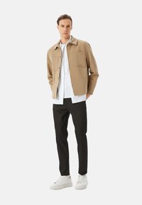 Veste beige à boutons, avec deux poches à l'avant, portée sur une chemise blanche, assortie à un pantalon noir et des baskets blanches. Design minimaliste.