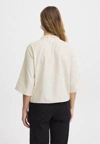 Krämfärgad blus med korta ärmar, vertikala blå ränder och volangkant vid halsringningen. Något kortare passform, slät tyg, minimalistisk design.