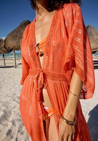 Cubre-bikini ligero y de color naranja, con abertura frontal, patrones intrincados y lazo en la cintura. Usado sobre un bikini multicolor. Fondo de playa de arena.