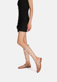 Sandali gladiator argento con lacci e suole piatte, caratterizzati da cinghie incrociate, punte aperte e un design minimalista.