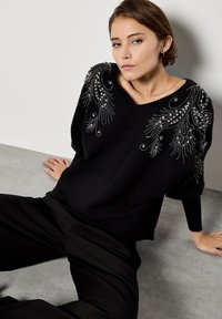 Czarny sweter dzianinowy z misternymi srebrnymi ozdobami na ramionach, z dekoltem w kształcie litery V i luźnym fasonem, w połączeniu z czarnymi spodniami.