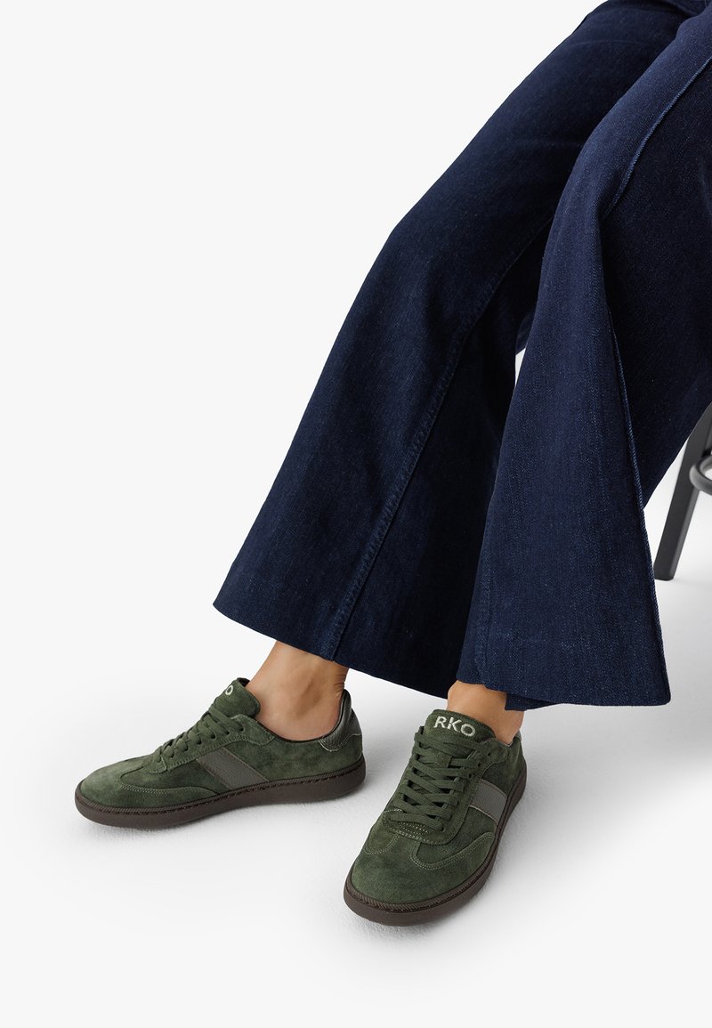 Jalkineet, joissa on vihreät mokkanahkakengät, joissa on "RKO" -merkintä kielen kohdalla, yhdistettynä tummansinisiin leveälahkeisiin farkkuihin, vaaleaa valkoista taustaa vasten.