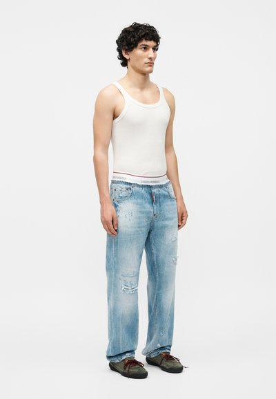 Νέος άνδρας με σγουρά μαλλιά που φοράει λευκό φανελάκι, ανοιχτό μπλε ταλαιπωρημένα τζιν και σκουροπράσινα αθλητικά παπούτσια, στέκεται μπροστά σε μονόχρωμο φόντο.